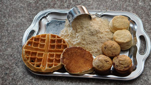 Silver tray on a granite counter holds waffles, pancakes, muffins, biscuits, and a measuring cup with flour. Cozy and inviting breakfast scene.