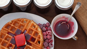 A waffle with red syrup and butter, surrounded by frozen raspberries, jars of preserves, and a bowl with a spoon on a brown tablecloth.
