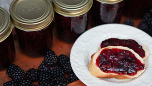 Jars of dark berry jam with gold lids are next to fresh blackberries. Two slices of bread with jam are on a white plate.