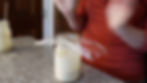 A person in an orange shirt gestures over a jar with plastic wrap on a granite counter. Another jar is nearby. Indoor kitchen setting.