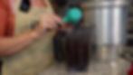 Person in apron using a teal scoop to add items to a blender, next to a large metal pot on a countertop. Kitchen setting.