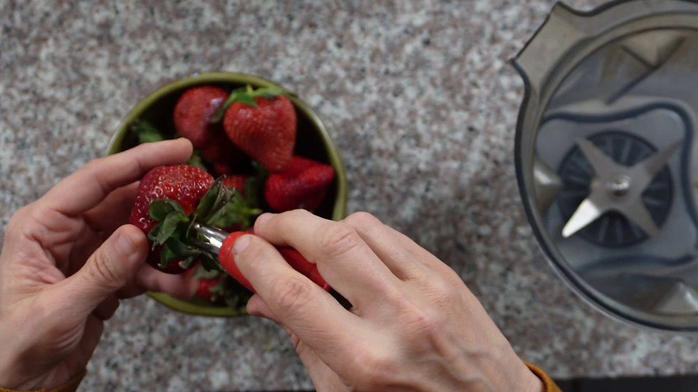 Hands hulling strawberries using a tool over a bowl on a granite countertop. A blender base is visible nearby.