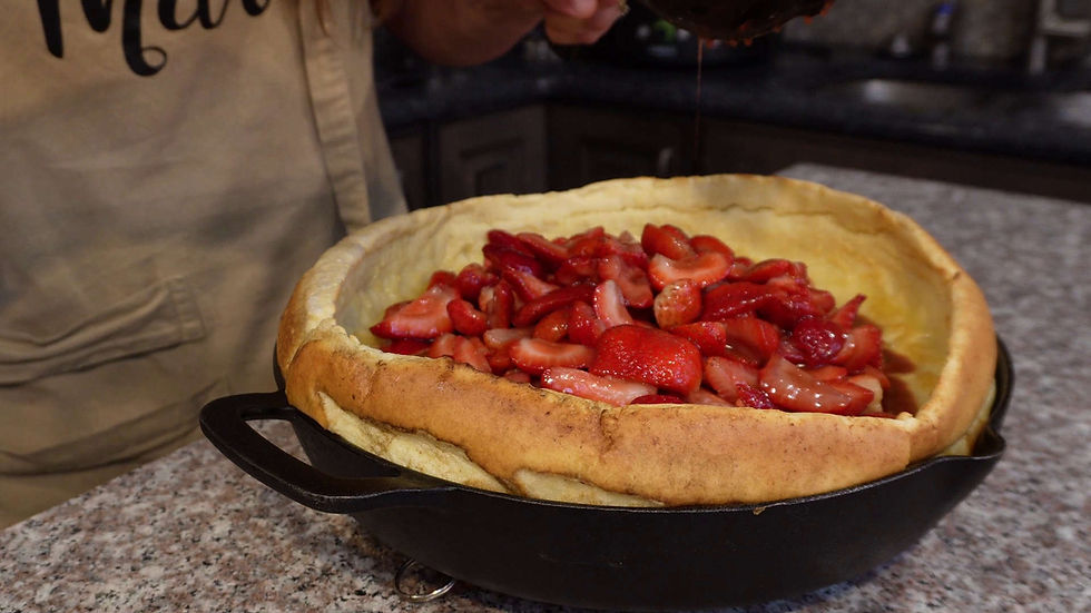 A person pours liquid over sliced strawberries in a large pancake in a skillet on a granite counter. A beige apron with "M" is partly visible.