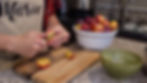 Person cutting pluots on a wooden board in a kitchen. A bowl of pluots and a green bowl are nearby. Apron text reads "Marie."