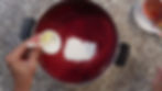 Hand adding butter to a pot of red liquid with white powder on a speckled countertop. A measuring spoon lies nearby.