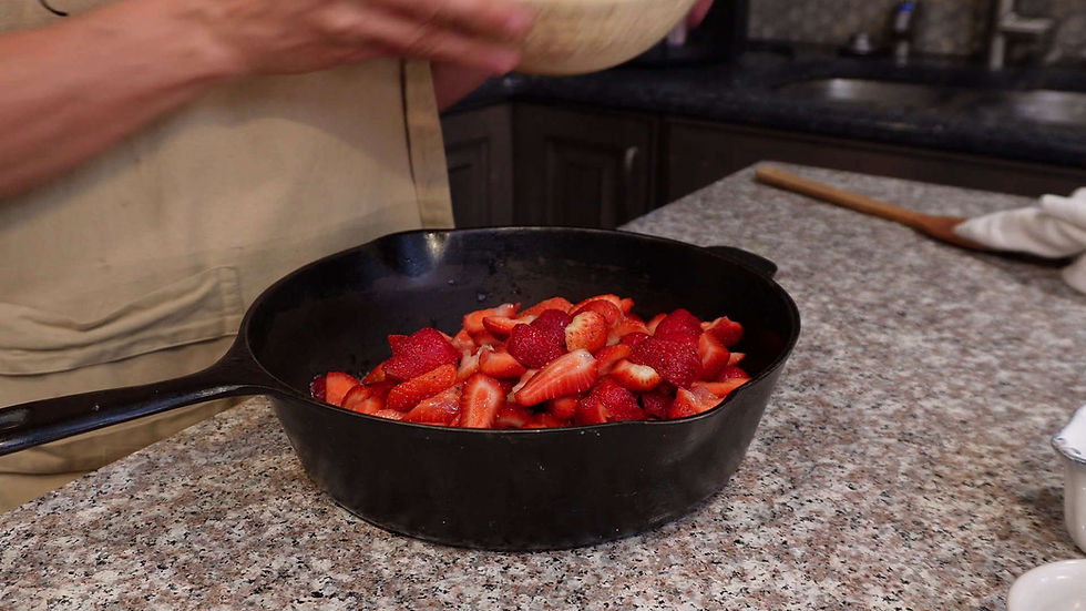 Person holding a bowl, adding strawberries to a black skillet on a granite countertop in a kitchen.