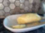 Butter on a white dish with a knife against a gray hexagonal-patterned tiled backsplash. Smooth, creamy texture displayed.