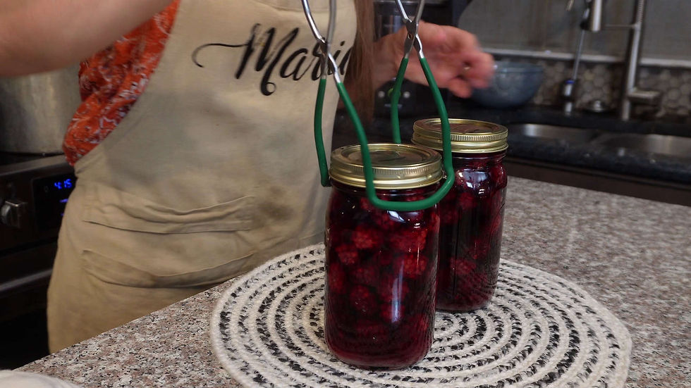 Person in a beige apron lifts jars of red berries with a jar lifter on a kitchen countertop. Apron text: "Marie". Brown and cream setting.
