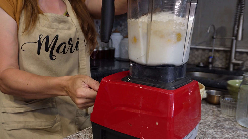 Person in a kitchen wears a beige apron labeled "Mari", using a red blender on a counter. The blender contains white liquid with yellow.