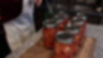 Person wearing apron uses tongs to handle jars of preserved fruit on a countertop. Kitchen sink in background, warm, homey atmosphere.