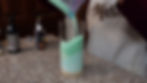 Person pours green sand from a bag into a tall glass on a marble counter. Bottles are nearby. Apron shows "Mar" text.