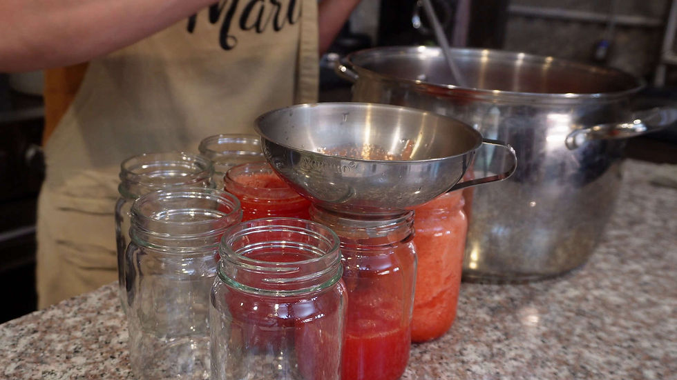 Canning jars being filled with tomato sauce using a funnel on a kitchen counter. A pot and a person wearing an apron are in the background.