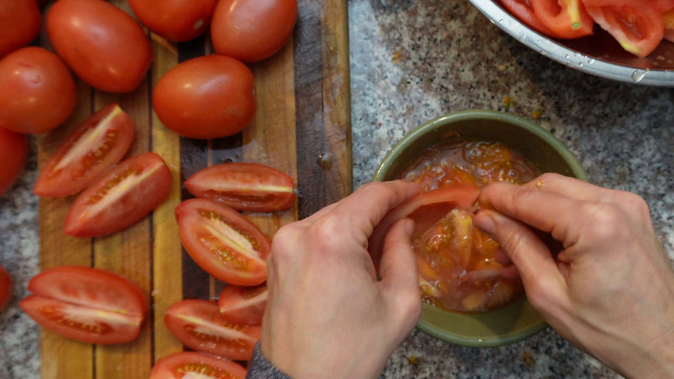 Hands deseeding tomatoes over a green bowl on a speckled countertop. Sliced tomatoes are on a wooden board beside whole ones.