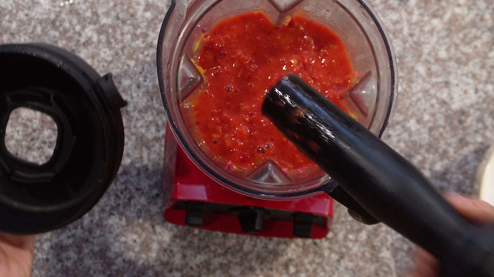 Red blender with an open lid, containing a thick red mixture. Black tamper in use on a speckled countertop. Hands hold the lid.