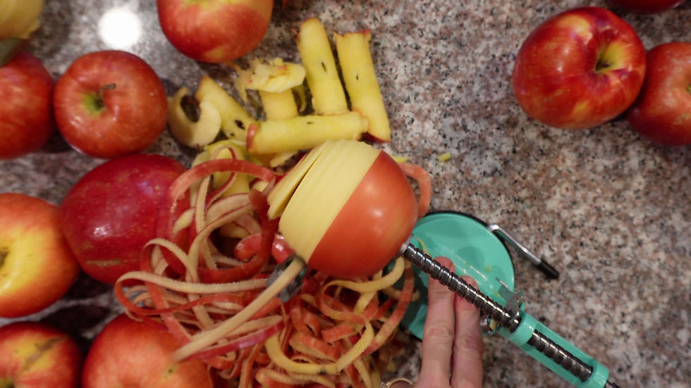 Hand using a teal apple peeler on a red apple, creating spiraled peels. Several whole apples and peeled cores on a granite countertop.