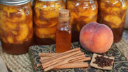Jars of preserved peaches, a glass bottle, a fresh peach, cinnamon sticks, and cloves on a patterned mat, evoking a warm, rustic feel.