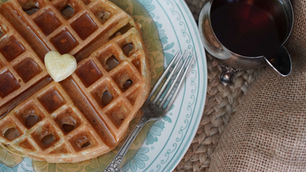 Waffles with syrup and heart-shaped butter on a floral plate, accompanied by a fork and a silver jug of syrup on a woven surface.