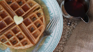 Waffles with syrup and heart-shaped butter on a floral plate, accompanied by a fork and a silver jug of syrup on a woven surface.
