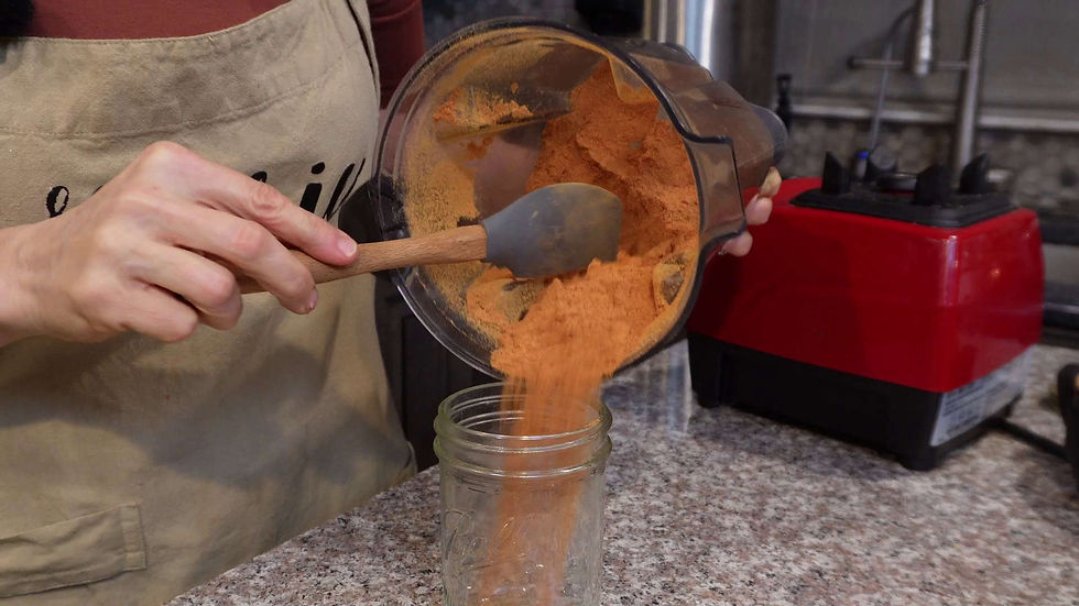 Person pouring orange powder from a blender into a jar on a granite counter. Wearing a tan apron with black text. Red blender in background.