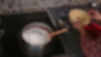 A pot on a stovetop with sugar and a wooden spoon. Hands hold a yellow bowl. Hexagonal tile backsplash and dark countertop visible.