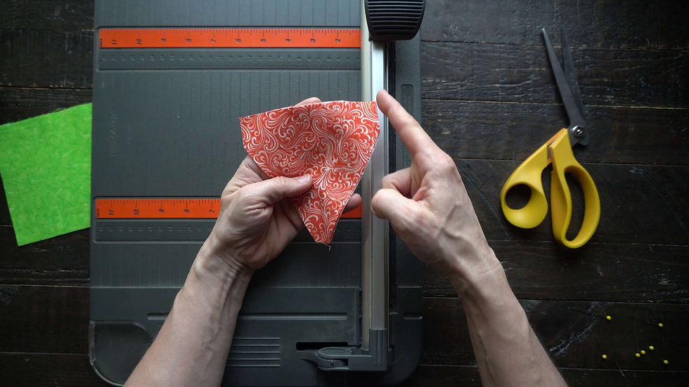 Hands folding an orange patterned paper near a paper cutter. Yellow scissors and green paper on a wooden table.