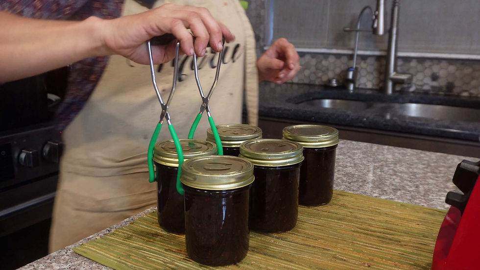 Person using tongs to handle jars of dark jam on a counter. Kitchen setting with sink and stove, beige apron visible. Calm atmosphere.