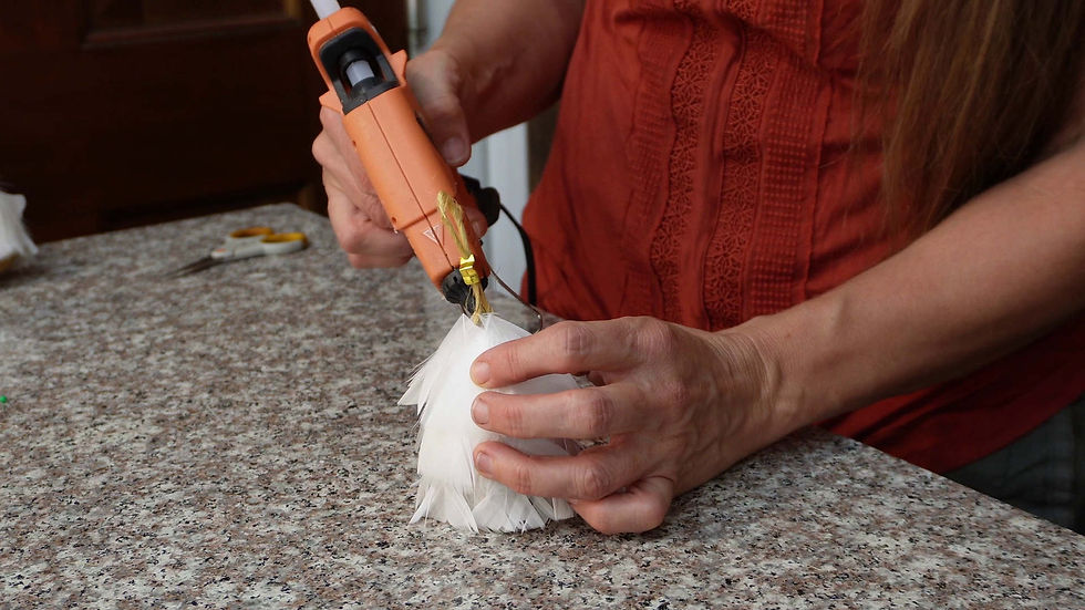 Hands using a hot glue gun to attach white feathers on a countertop. Person wearing an orange textured top, scissors visible in the background.