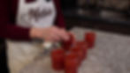 Woman in a maroon sweater and apron labeled "Marie" cleans tops of six red jam jars on a granite counter in a kitchen setting.