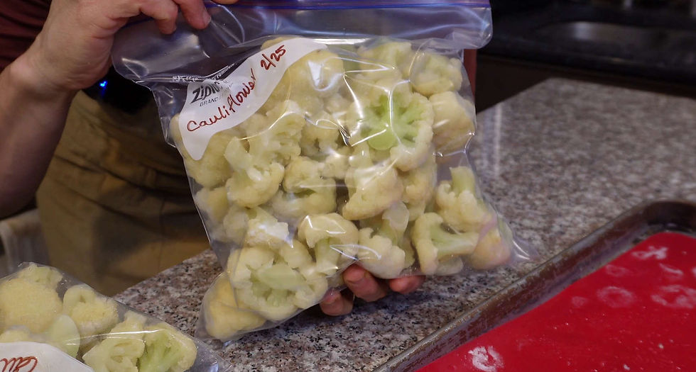 Hand holds a clear bag of frozen cauliflower labeled "Cauliflower 2/23" on a granite countertop. Red mat visible at the bottom.