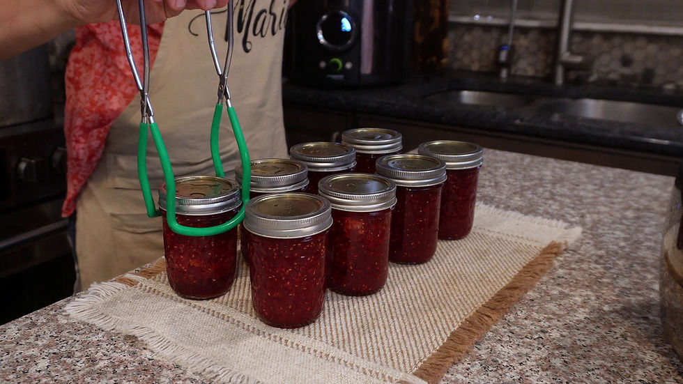 Person using tongs to move jars of red jam on a countertop. Apron says "Marie." Kitchen background with patterned counter and cloth.