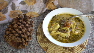Bowl of tortellini soup with herbs and meat on woven mat. A pinecone and sunflower-patterned cloth nearby, on a speckled countertop.