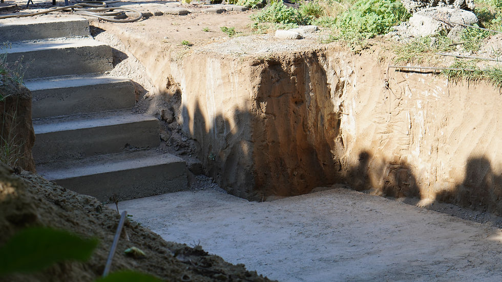 Excavated trench with concrete steps at a construction site, sunlit dirt walls and scattered grass.