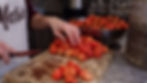 A person prepares strawberries on a wooden board, removing stems with a knife. A large metal bowl of strawberries is in the background.