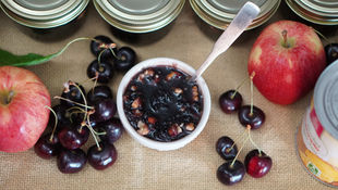 Bowl of dark fruit jam with a spoon, surrounded by cherries, apples, and canned goods on burlap. Jars with metal lids in the background.