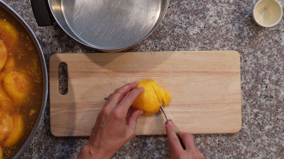 Hands slicing a peeled peach on a wooden board, set on a speckled countertop. Nearby, peaches are in a bowl, with a metal pan and small cup.