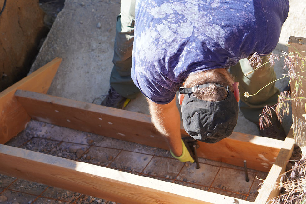 Worker in cap and gloves bending over a wooden frame, fastening rebar in a sunny construction site.