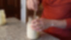 Hands stirring creamy mixture in a mason jar on a granite countertop. Person wears an orange shirt, showing engagement ring. Warm lighting.