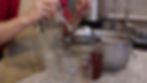 A person in a red shirt and beige apron uses a ladle to pour liquid into jars in a kitchen. The apron has text "Mari...". A pot is visible.