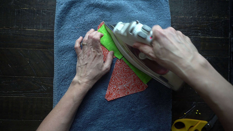 Hands ironing red and green fabric on a blue towel. Yellow scissors and pins on wood surface suggest a sewing project.