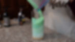 Person pours green powder into a glass on a granite countertop. Bottles and a text apron in the background. Creative and focused setting.