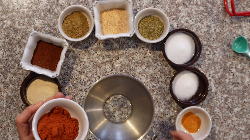 Various colorful spices in white bowls arranged on a speckled countertop. A hand holds a bowl of red spice, creating a vibrant scene.