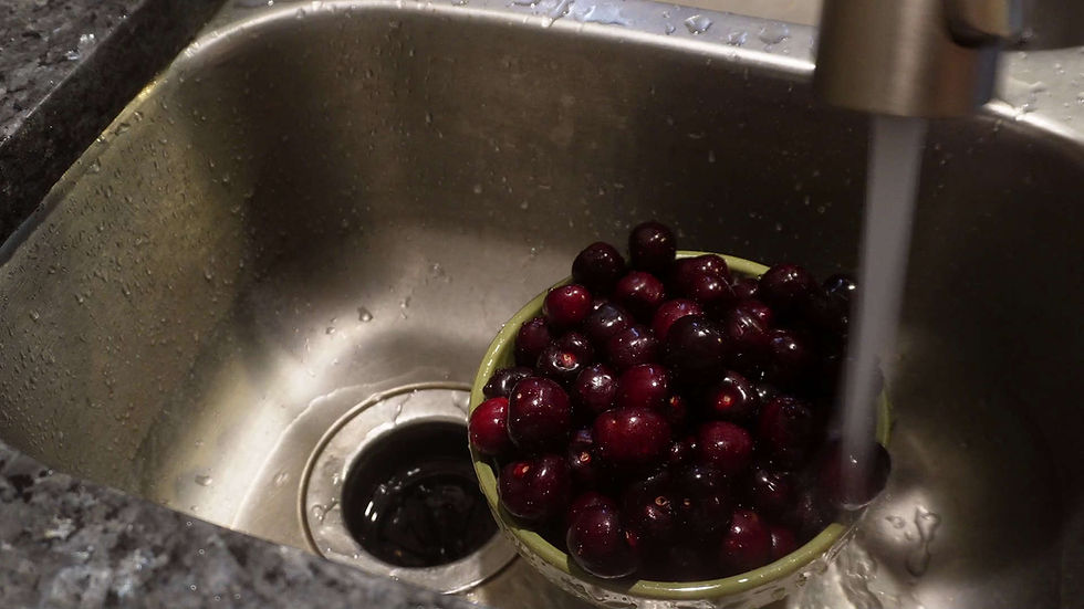 A colander of dark cherries being rinsed under running water in a stainless steel sink. Water droplets are visible on the sink's surface.