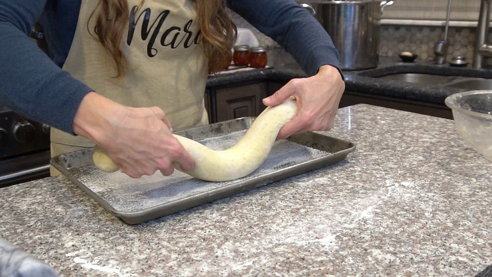 Person stretching dough on a floured baking sheet in a kitchen. Wearing an apron with "Maria" text. Countertop and jars in background.