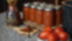 Jars of tomato sauce on a wooden board, next to ripe tomatoes and a ceramic jug, with a heap of spice on a spoon. Warm, rustic kitchen vibe.