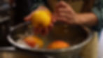 Hands peeling peaches in a metal bowl filled with iced water, in a kitchen setting. Peaches are orange and yellow. Striped sleeve visible.
