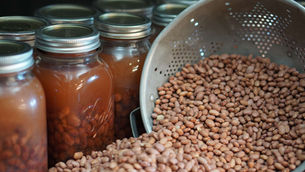 Jars filled with preserved beans sit next to a colander full of beans. The setting is a kitchen with a focus on home canning.