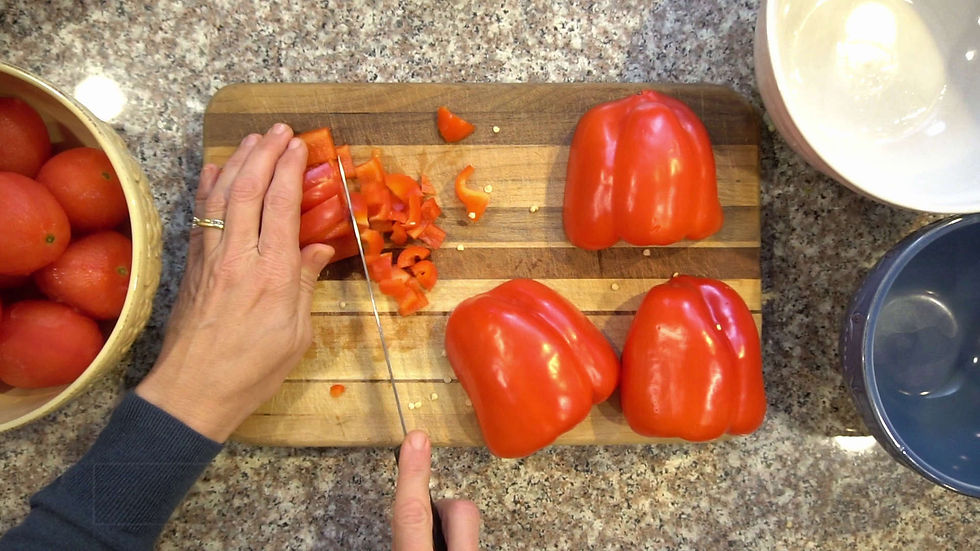Hands chopping red bell peppers on a wooden board, surrounded by tomatoes. Background shows a granite countertop and bowls.
