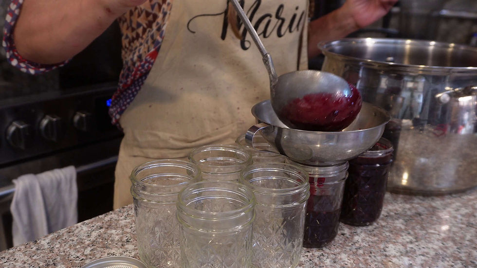Person ladling purple jam into jars in a kitchen. Apron with "Marie" text. Granite countertop, patterned shirt, and stainless steel pot visible.