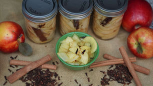 Jars of sliced apples with cinnamon, a green bowl of apple slices, red apples, cinnamon sticks, and cloves on burlap.