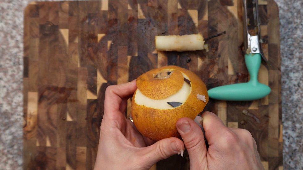 Hands peeling a pear with a vegetable peeler on a wooden cutting board. A pear core and teal peeler are on the board.